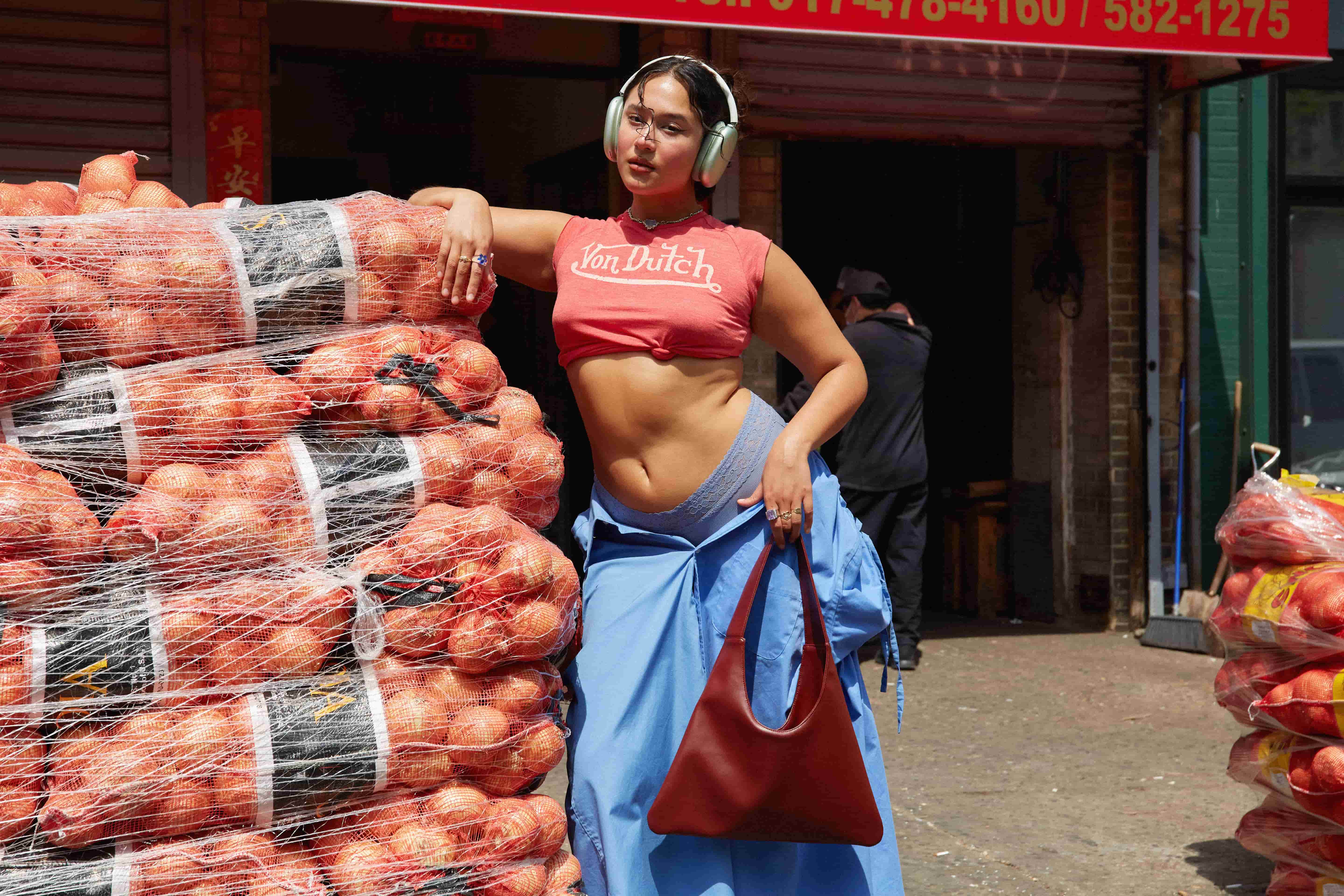 Woman leaning on a stack of onions with a red shirt and blue skirt and a sustainable red leather handbag made from cactus leather in an outdoor setting.