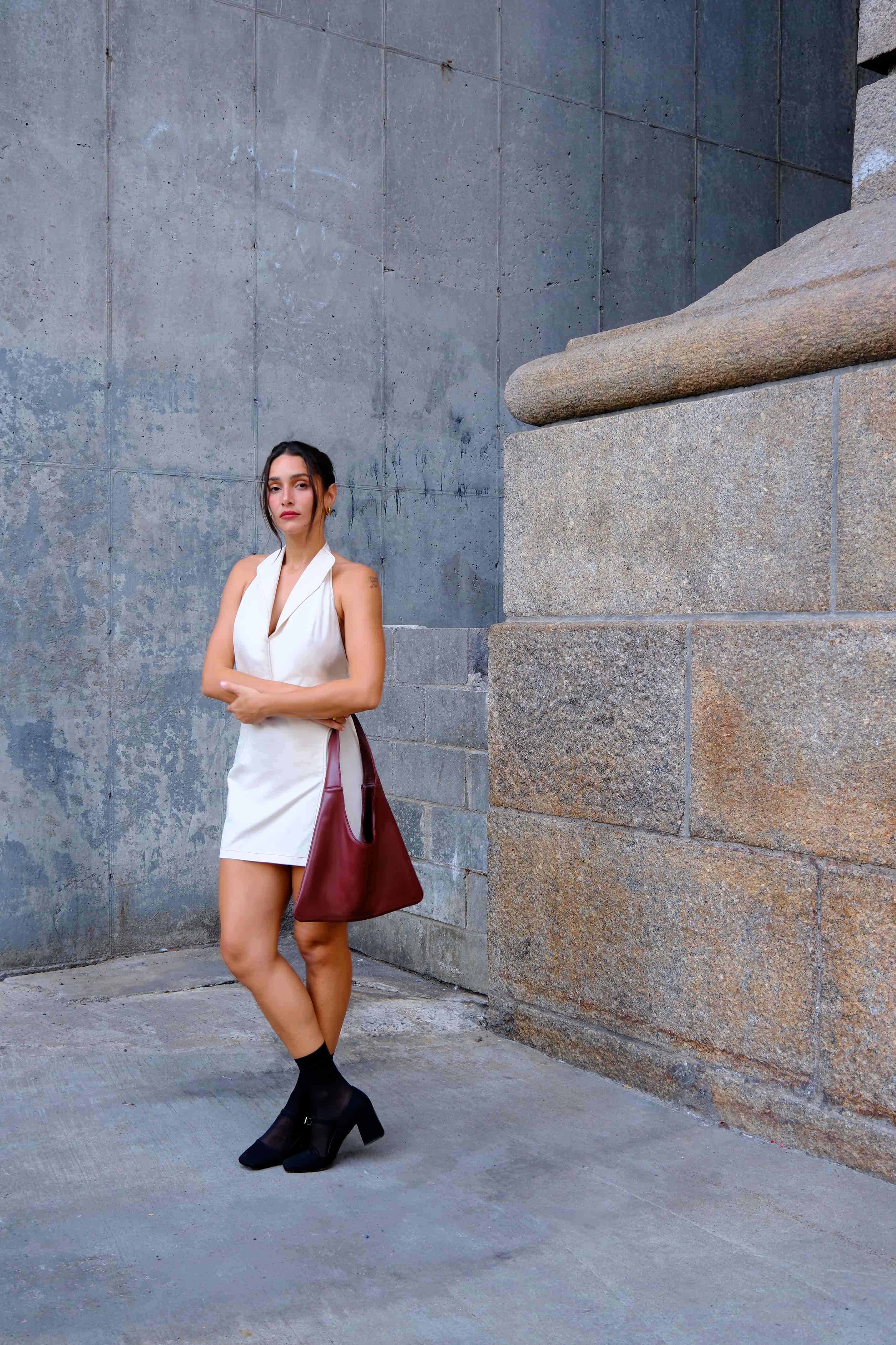 Woman in a white dress with a red sustainable tote bag made from cactus leather standing against a stone wall.