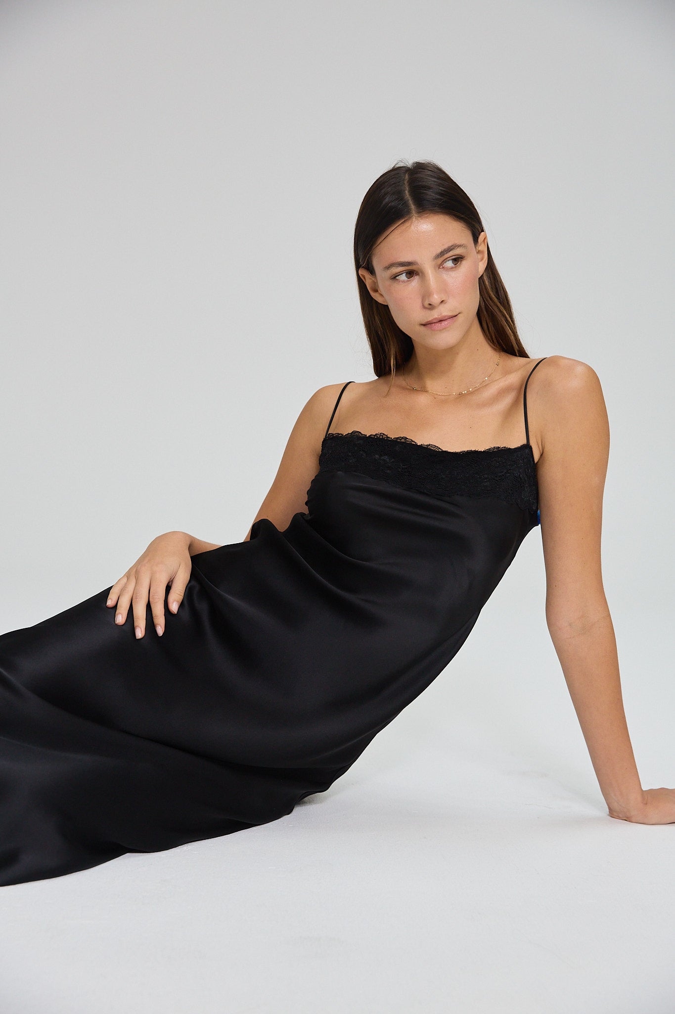 Model wearing a black, midi-length dress with two shoulder strap, featuring lace detail in a white background. Made from sustainable materials.