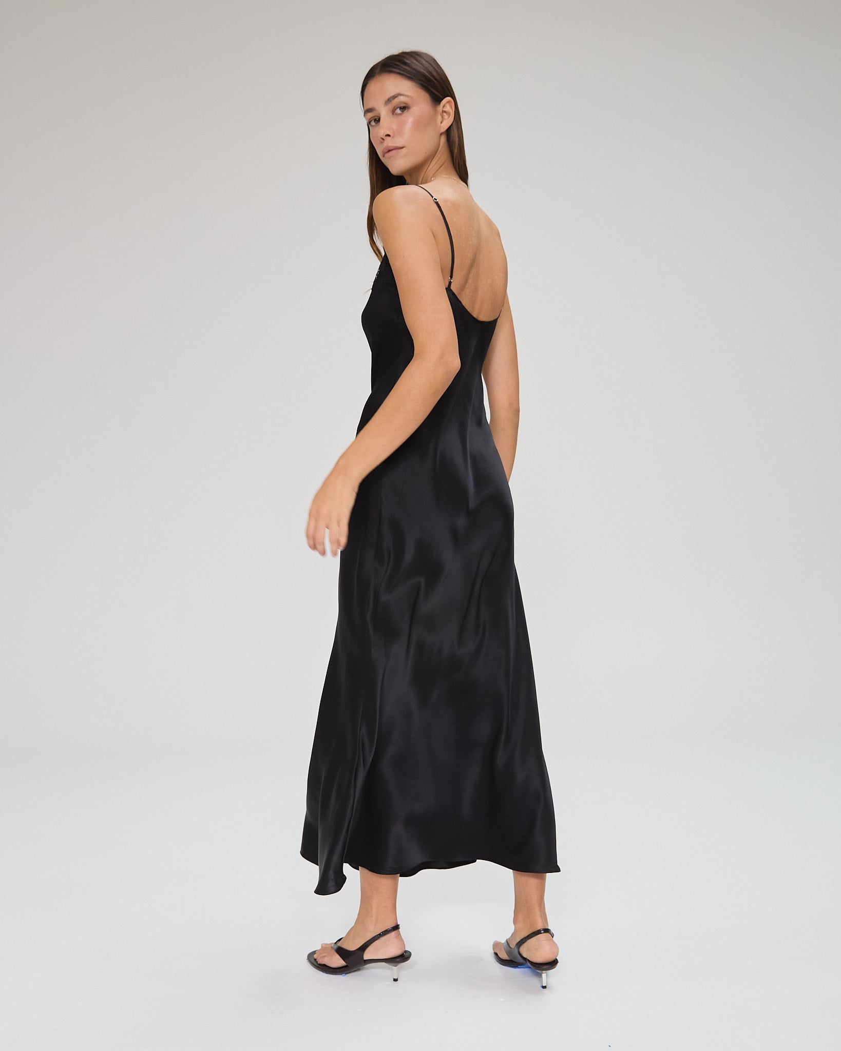 Model wearing a black, midi-length dress with two shoulder strap, featuring lace detail in a white background. Made from sustainable materials.