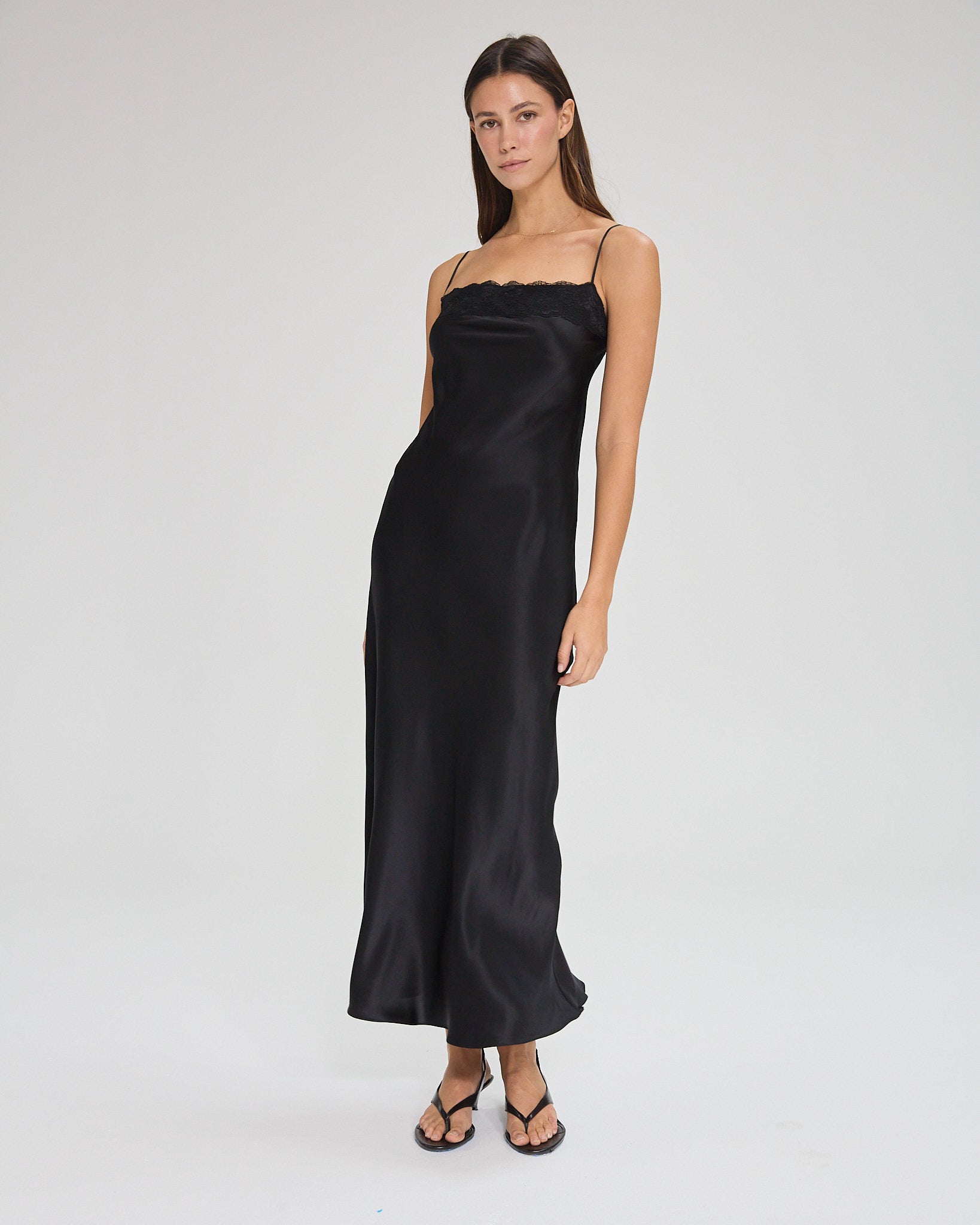 Model wearing a black, midi-length dress with two shoulder strap, featuring lace detail in a white background. Made from sustainable materials.