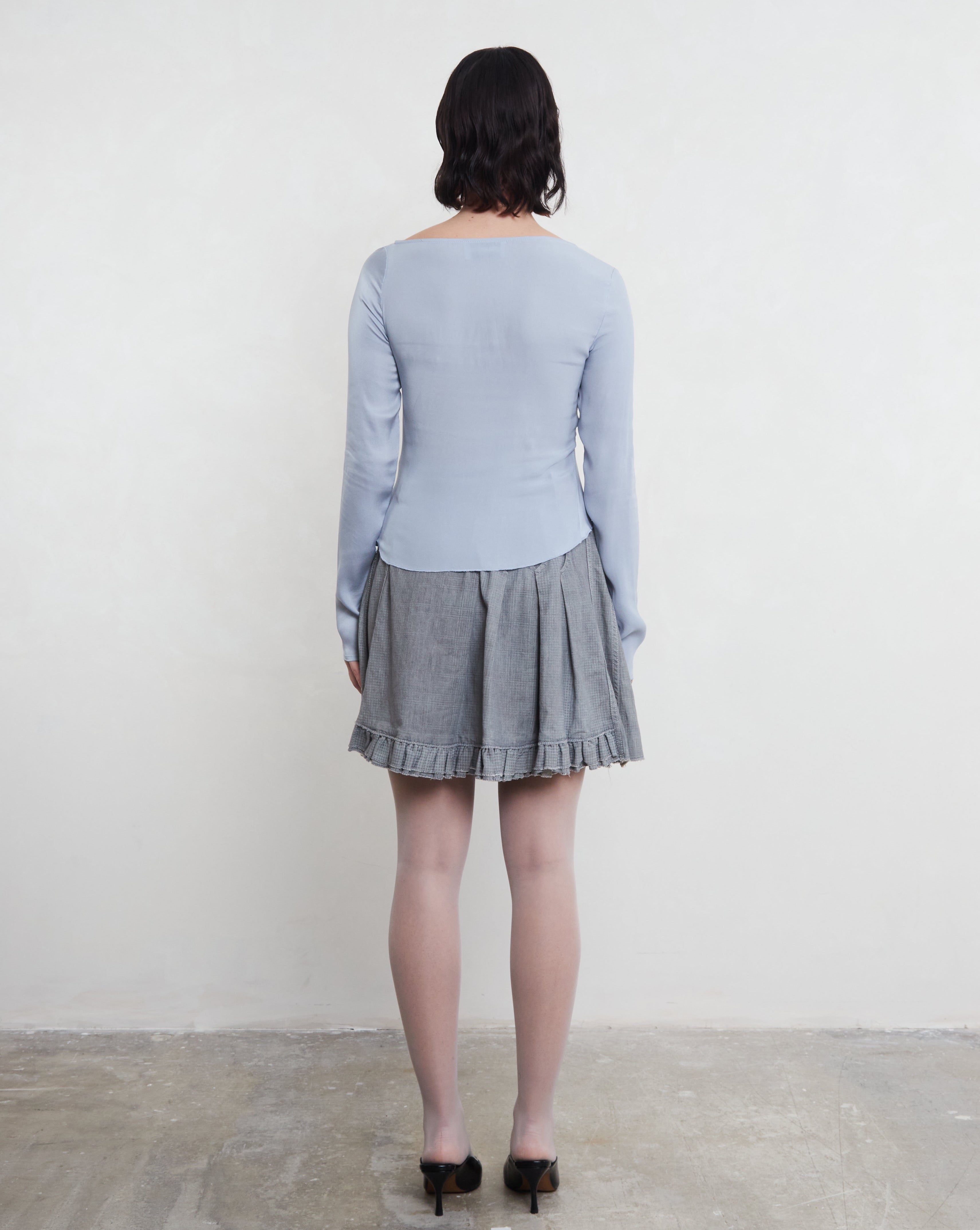 Woman wearing a light blue sustainable silk top and gray pleated skirt against a plain background.