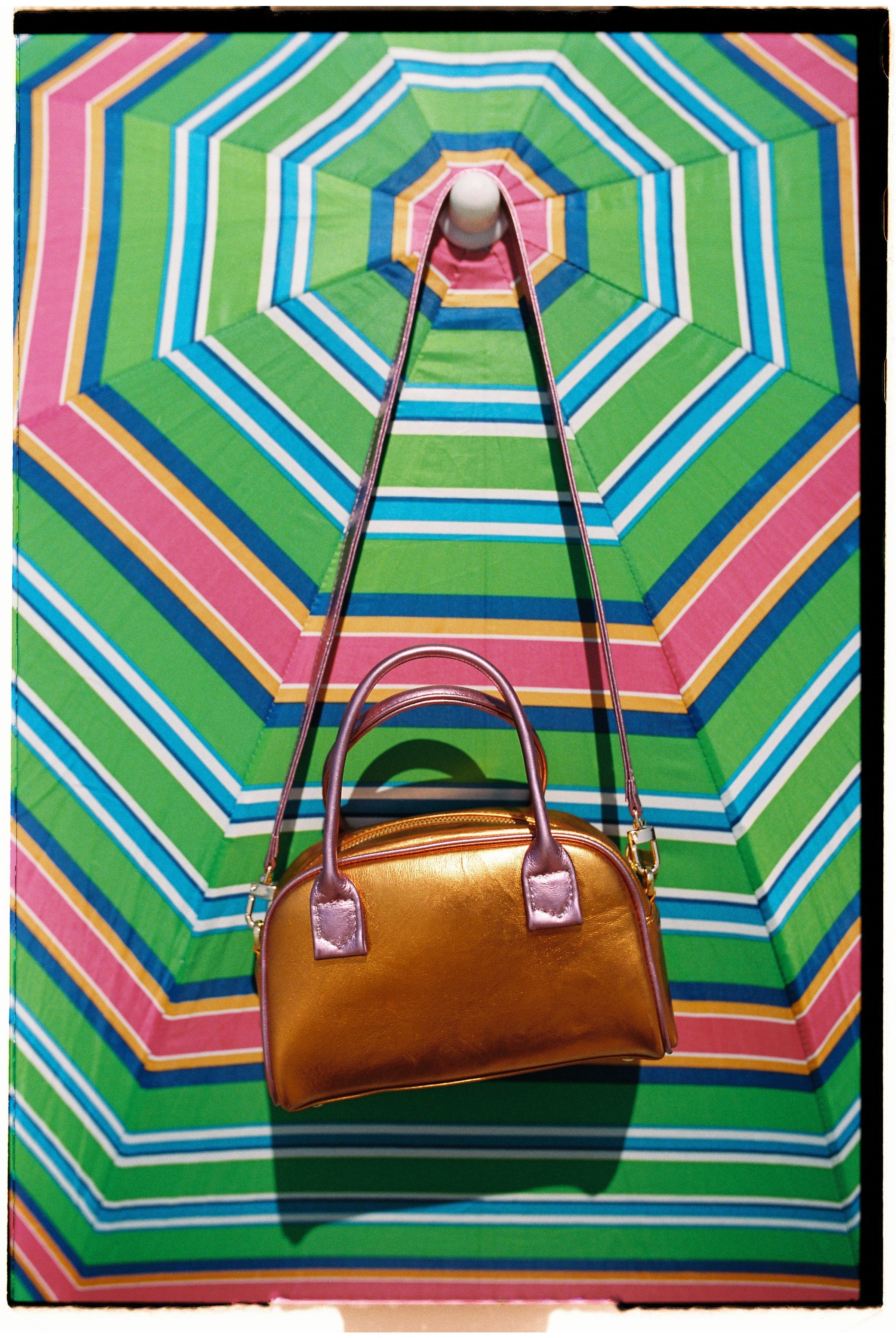 An orange and light pink handbag with a removable strap displayed hanging on a striped umbrella. 100% deadstock leather.