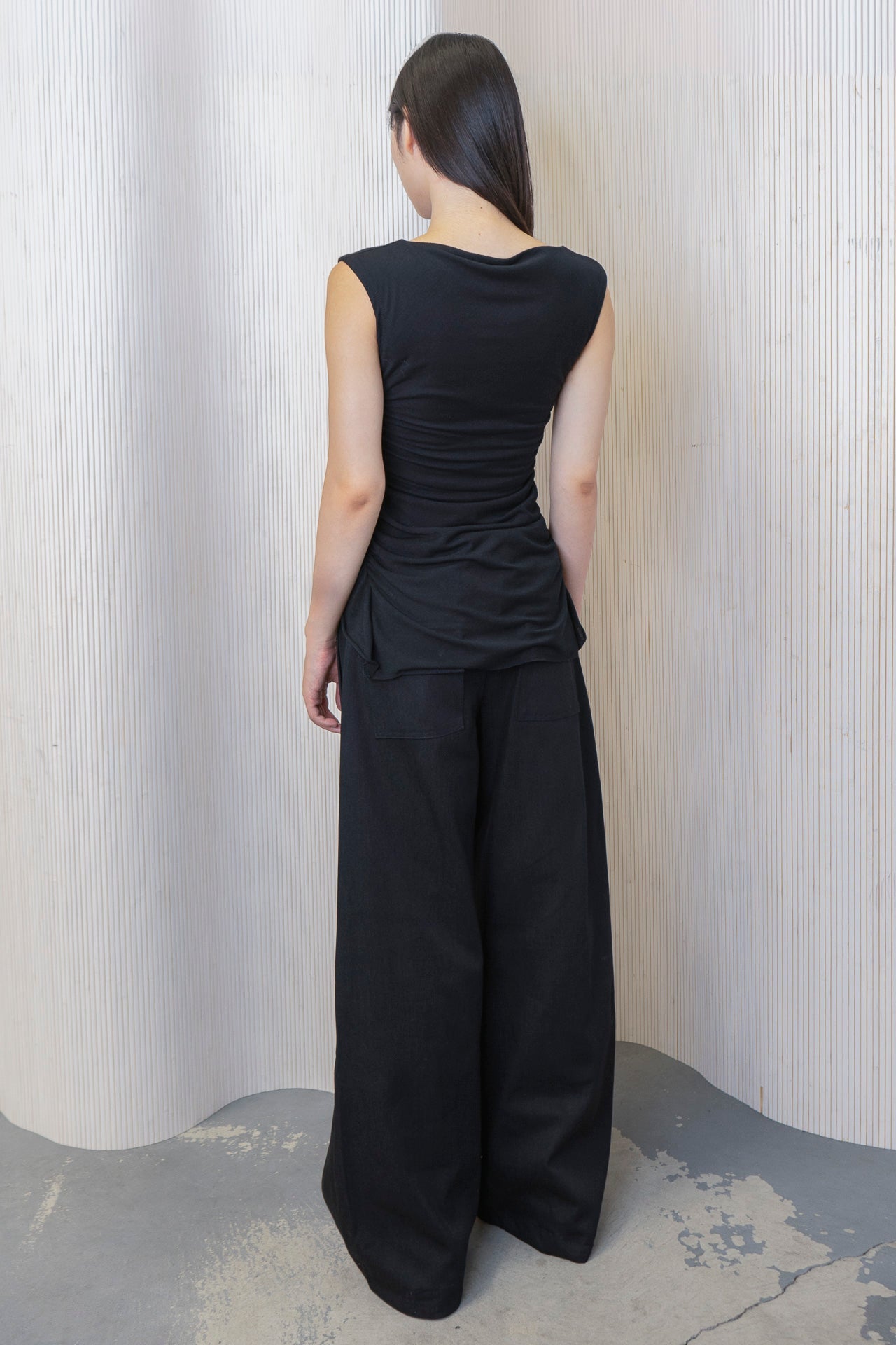 A woman wearing a black boat-neck top with side ruching and a high-low hemline, paired with black trousers and a logo belt. Made from deadstock fabric.