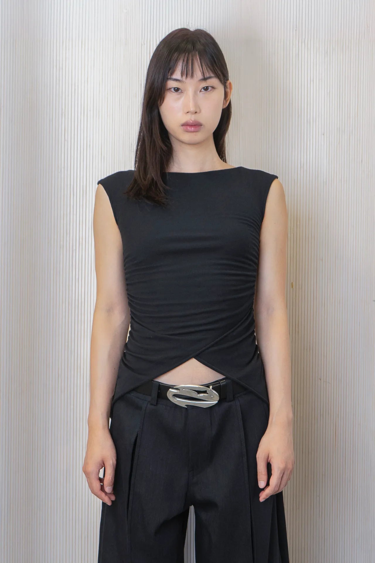 A woman wearing a black boat-neck top with side ruching and a high-low hemline, paired with black trousers and a logo belt. Made from deadstock fabric.