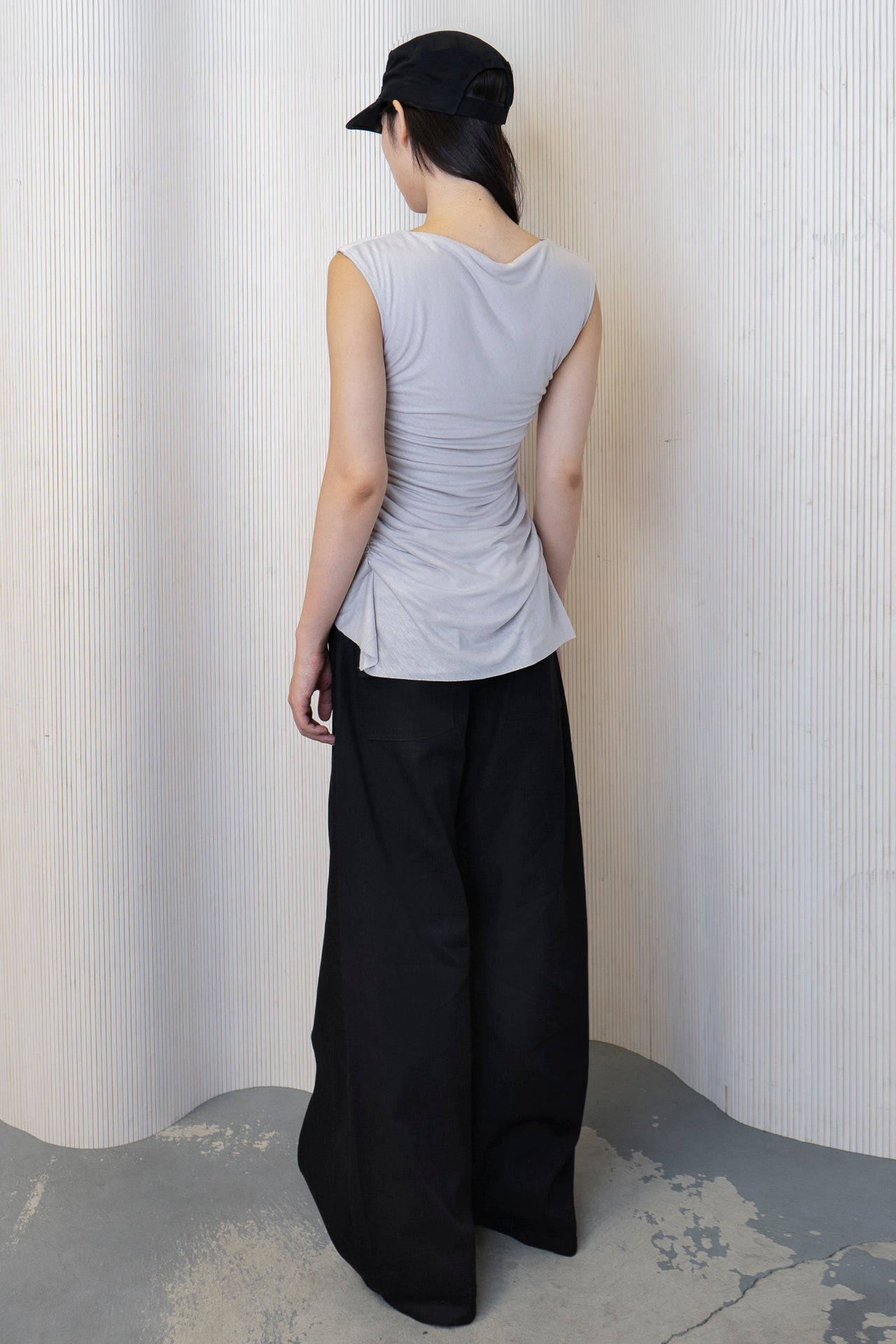 A model wearing a boat-neck top with a subtle back cowl neckline, side ruching, and a high-low hemline in a bone color. Made from 100% deadstock fabric. Back view.