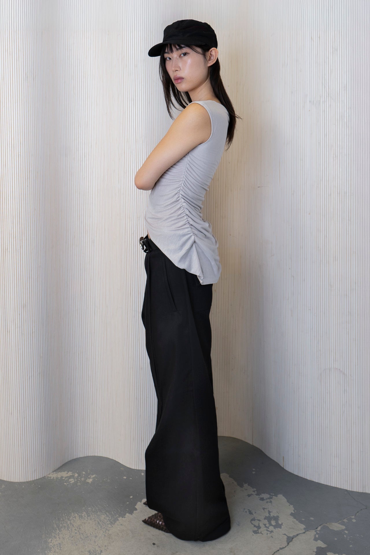 A model wearing a boat-neck top with a subtle back cowl neckline, side ruching, and a high-low hemline in a bone color. Made from 100% deadstock fabric. Side View.