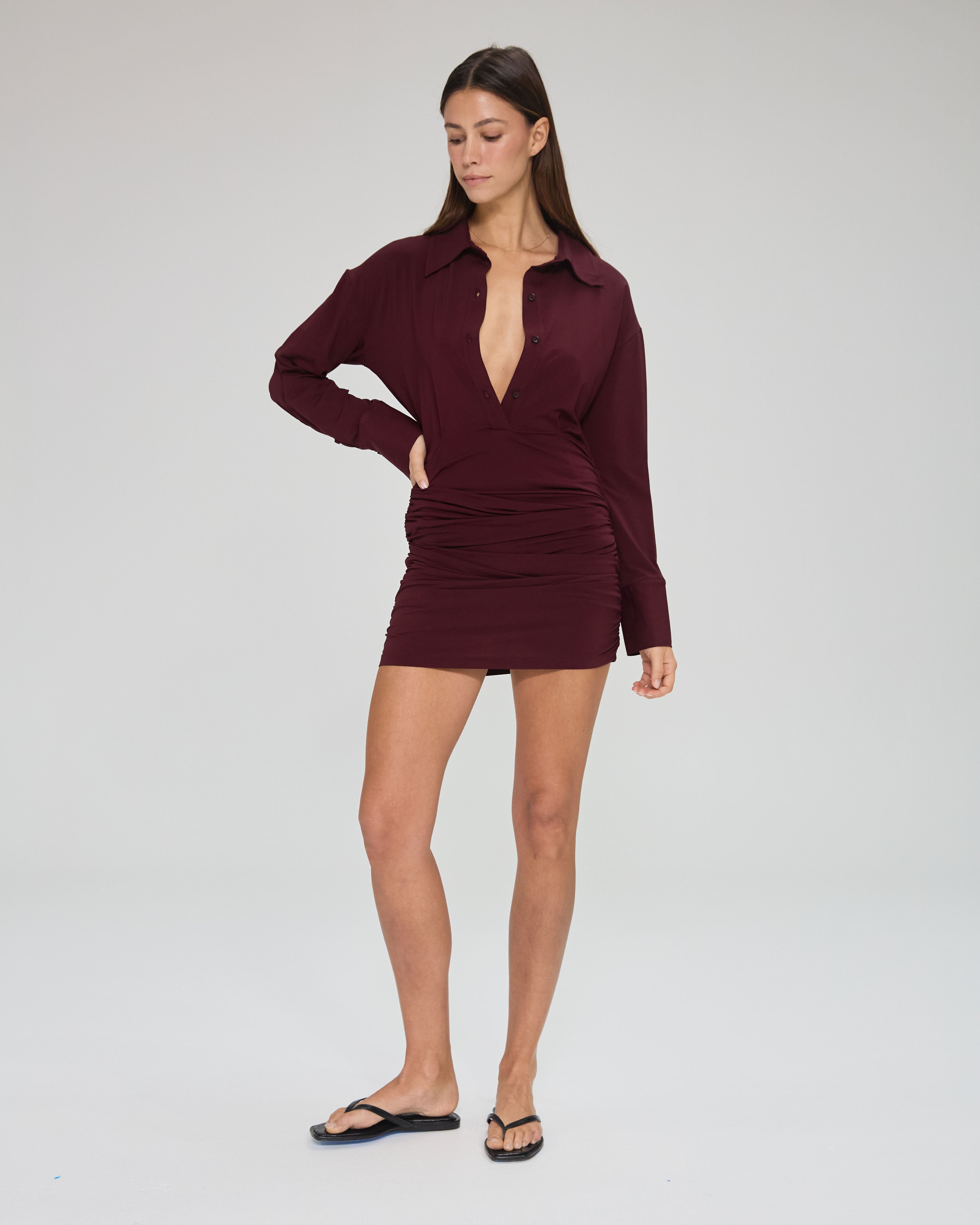 Model wearing a burgundy, mini-length long sleeve dress with buttons and collar in a white background. Made from sustainable deadstock materials.