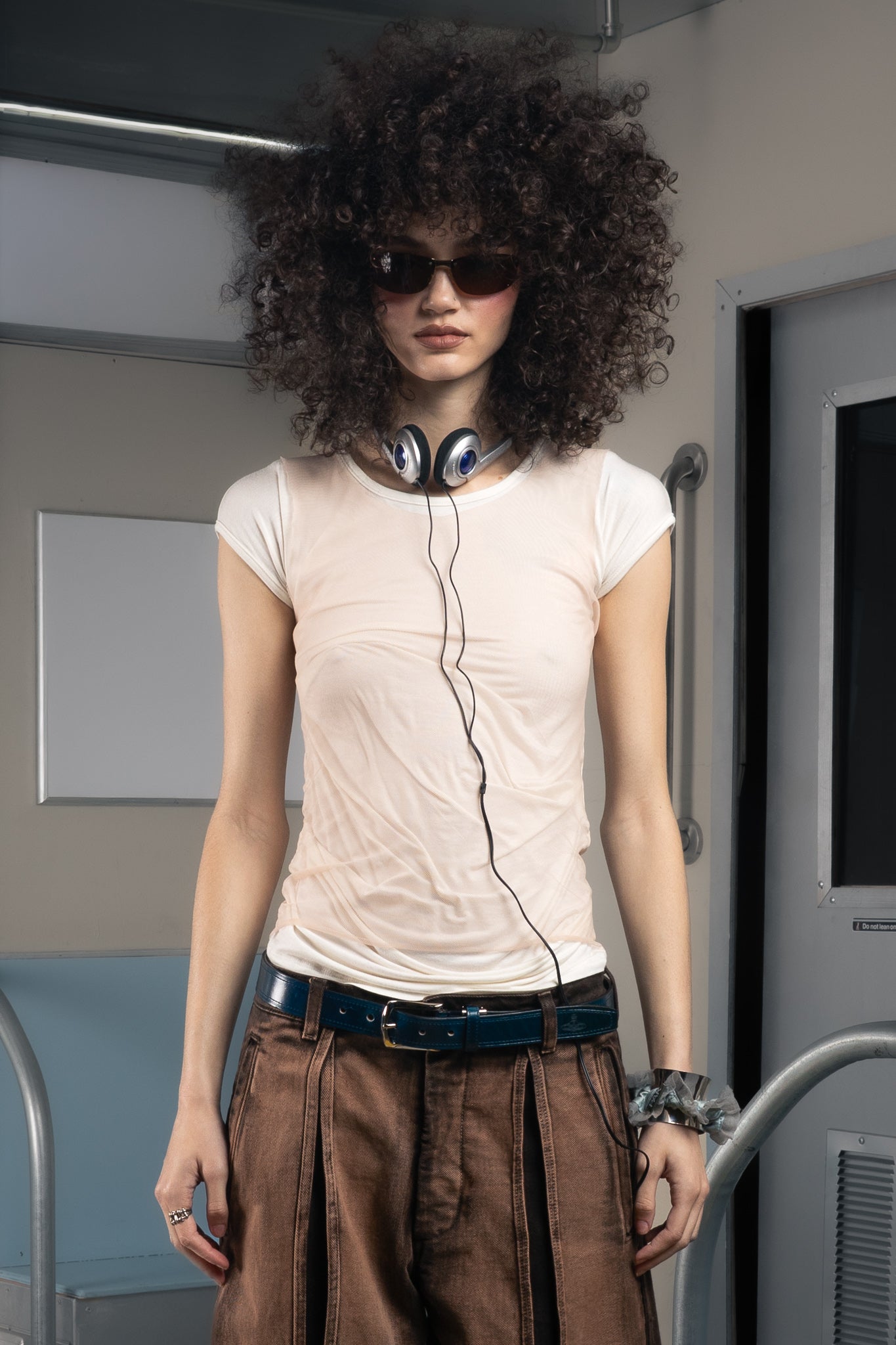 Person with an afro hairstyle wearing sunglasses and a light pink sustainable t-shirt inside a subway.