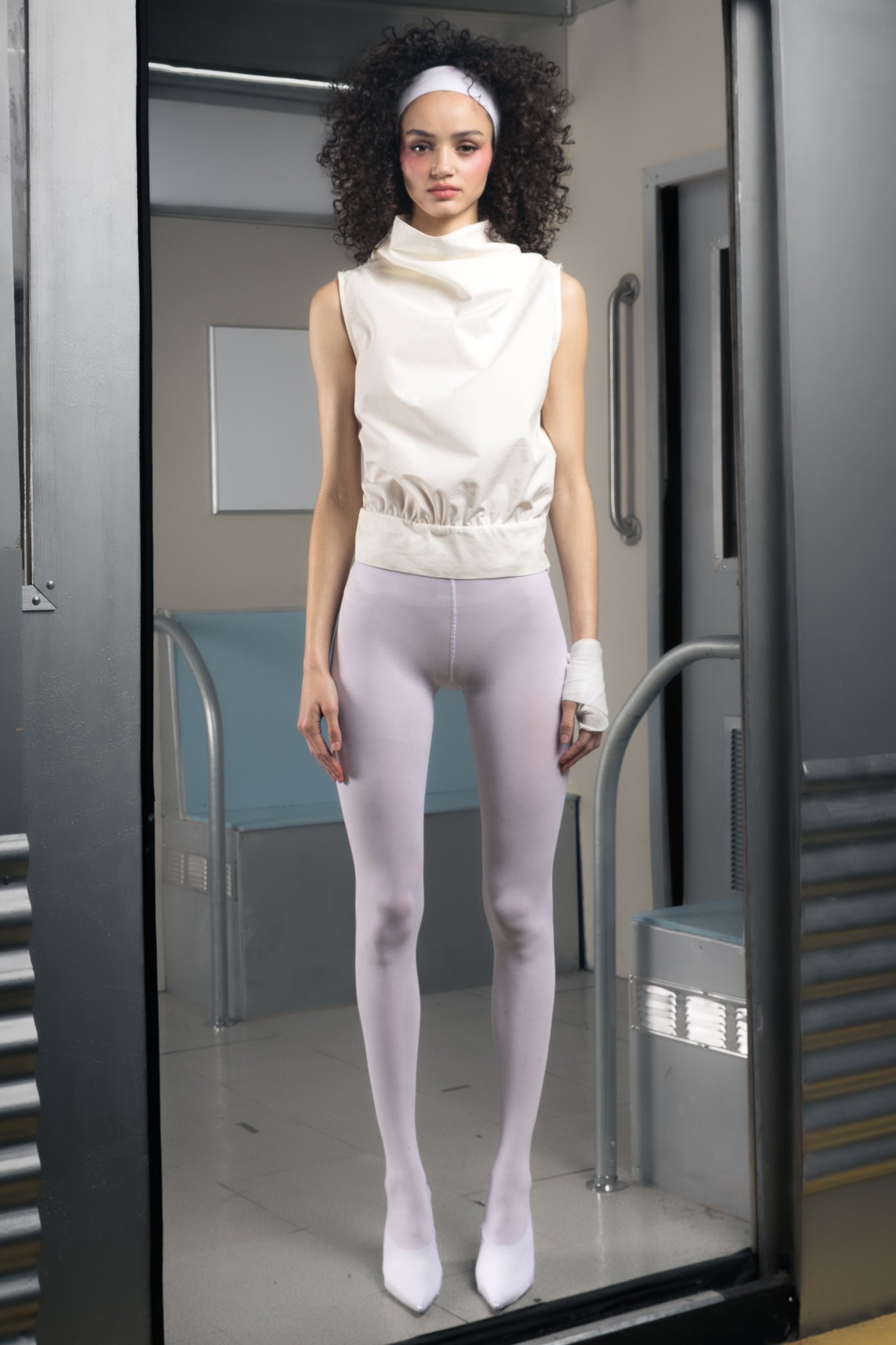 Woman wearing a white sustainable sleeveless top and tights in a room with metallic walls.