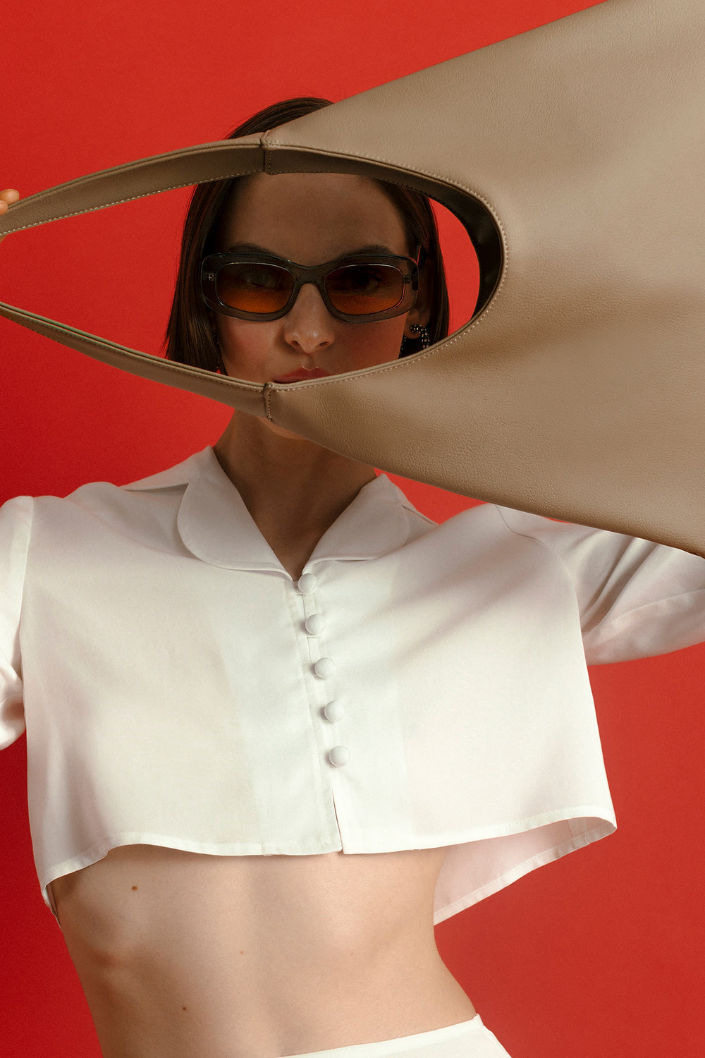Model wearing a white shirt and sunglasses holding a beige bag made from cactus leather and 100% organic cotton. On a white background against a red background. 