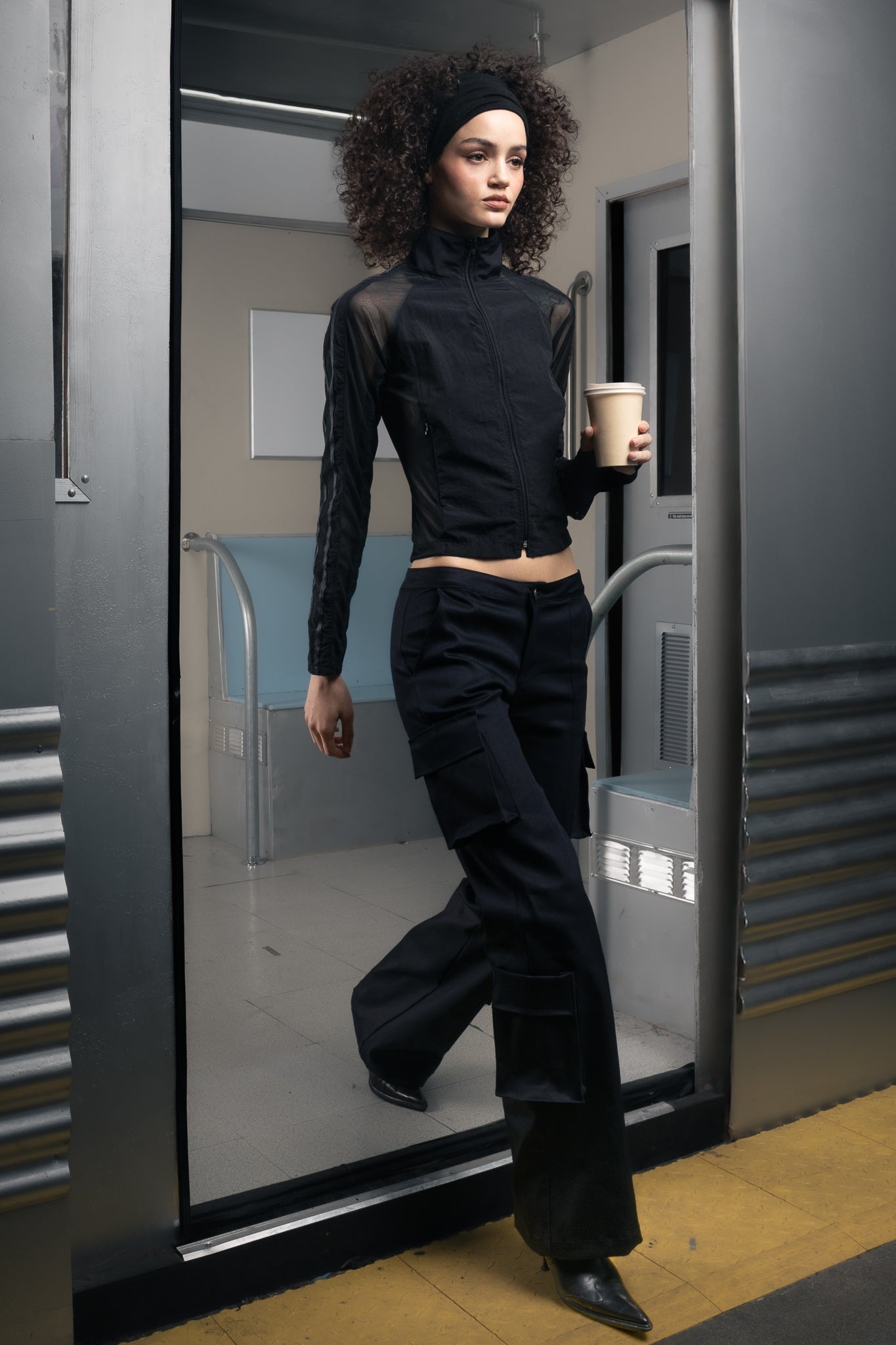 Woman in a sustainable black outfit holding a coffee cup and walking out of a futuristic train.