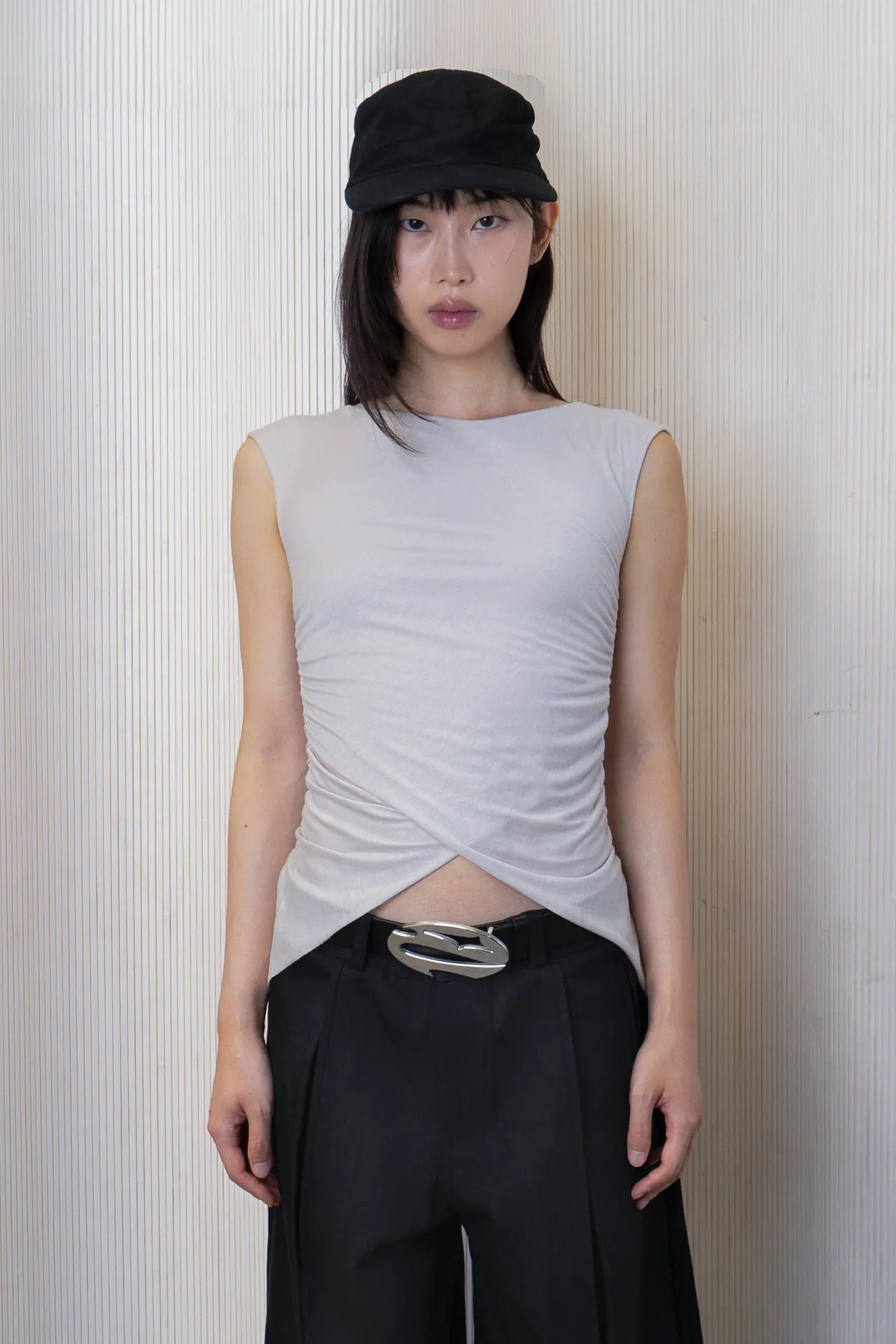 A woman wearing a sleeveless boat-neck top with side ruching and a high-low hemline, paired with black trousers and a belt. Made from 100% deadstock fabric. 