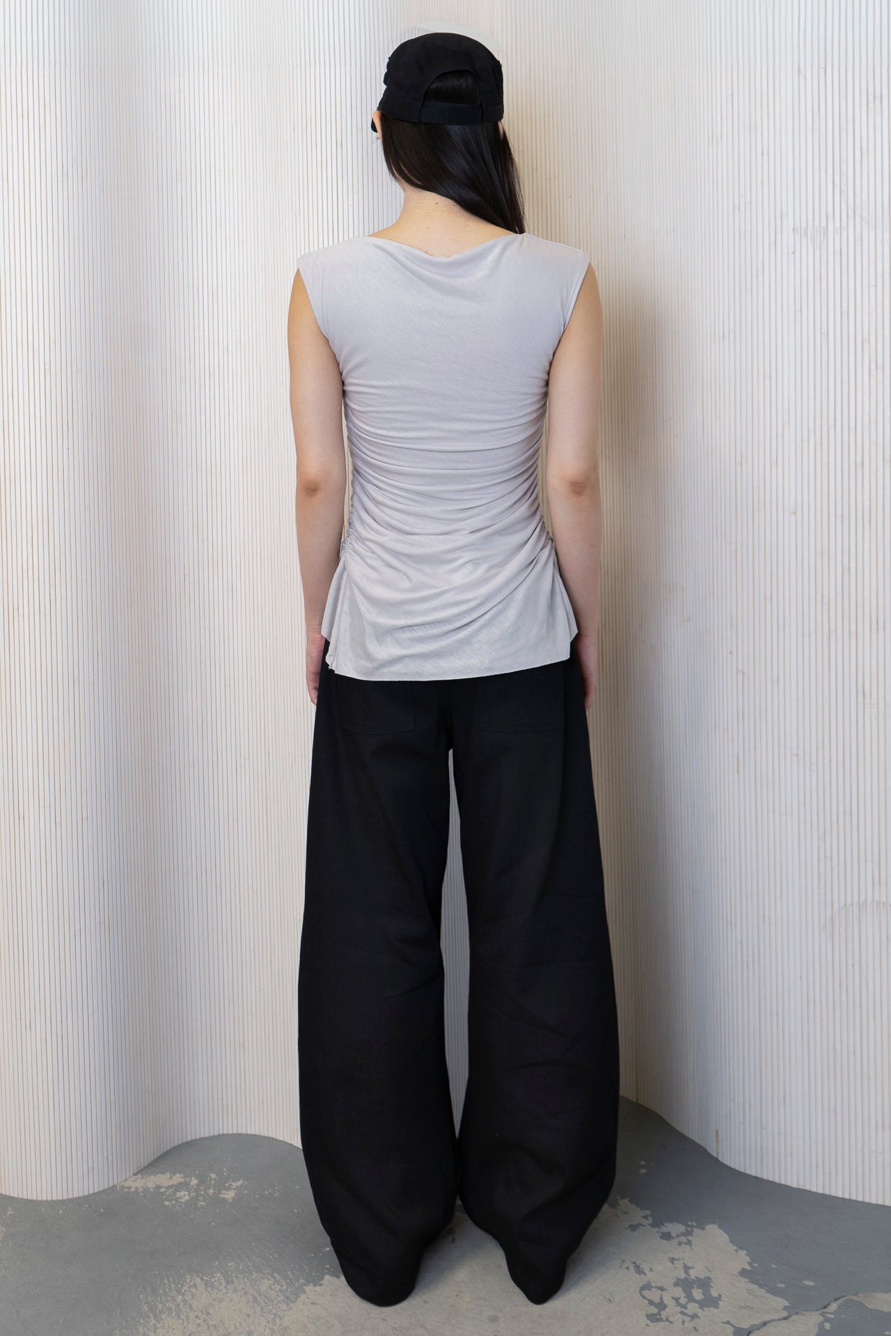 A model wearing a boat-neck top with a subtle back cowl neckline, side ruching, and a high-low hemline in a bone color. Made from 100% deadstock fabric. Back View.